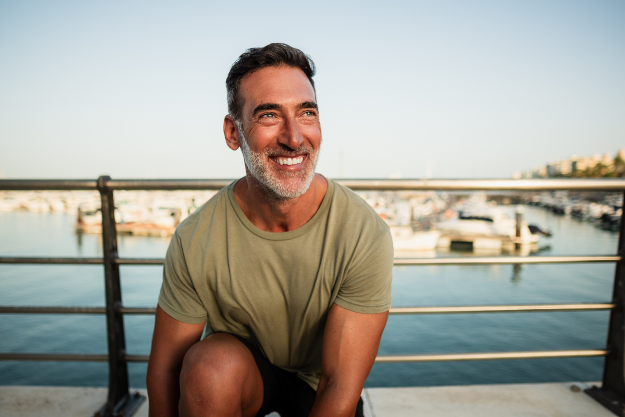 Portrait of a happy mature man smiling and kneeling by the sea, enjoying a sunny day